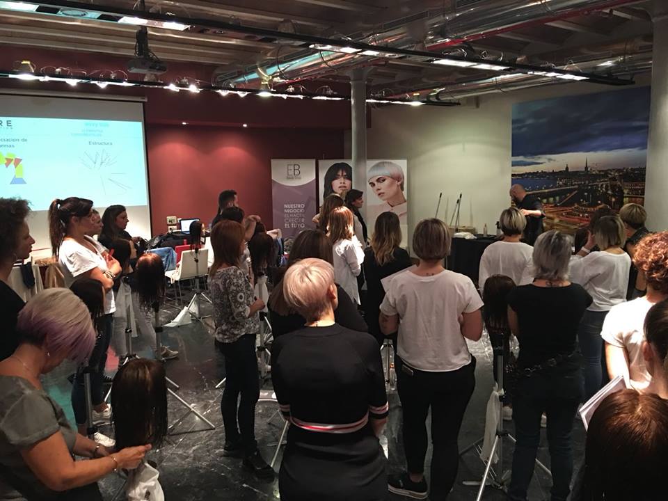 Clase de peluquería: un grupo de estilistas observa una demostración. Maniquíes, pantalla de presentación y pared roja.