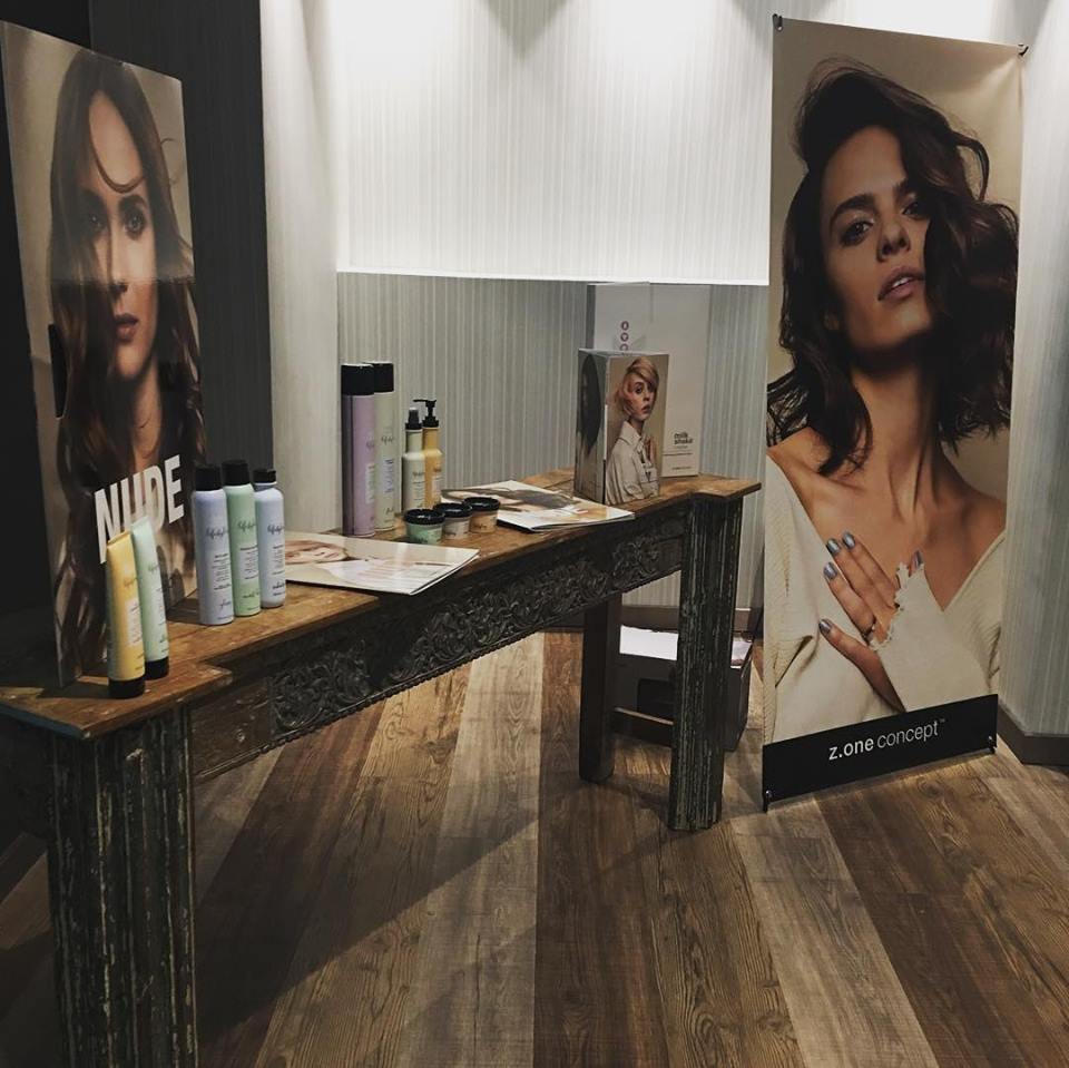 Exhibición de salón: productos sobre una mesa rústica con pancartas promocionales que presentan mujeres con cabello peinado.