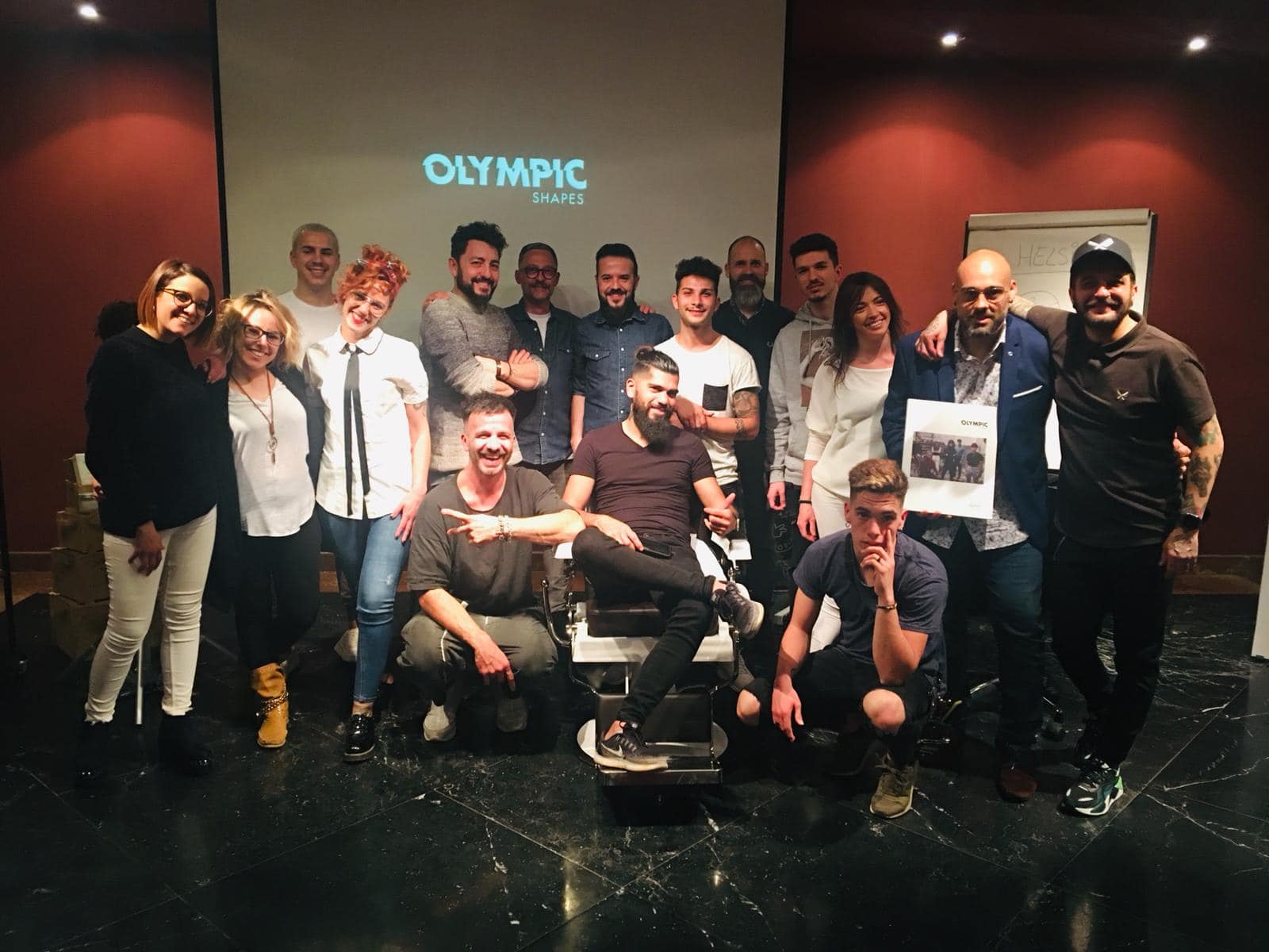 Foto grupal en una sala con el logo olímpico en pantalla. Personas sonriendo, algunas con un premio.