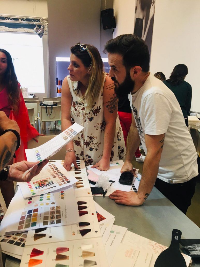 Personas revisando muestras de diseño en una mesa: mujer con vestido floral, hombre con barba, papeles y muestras de color.