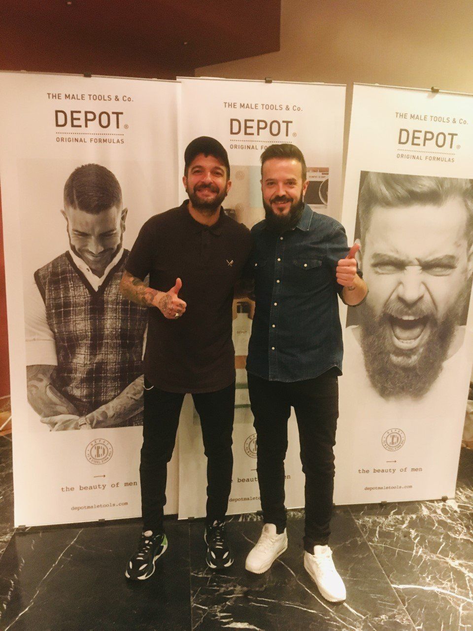 Dos hombres sonriendo, con los pulgares hacia arriba, frente a la pancarta del Depósitos.