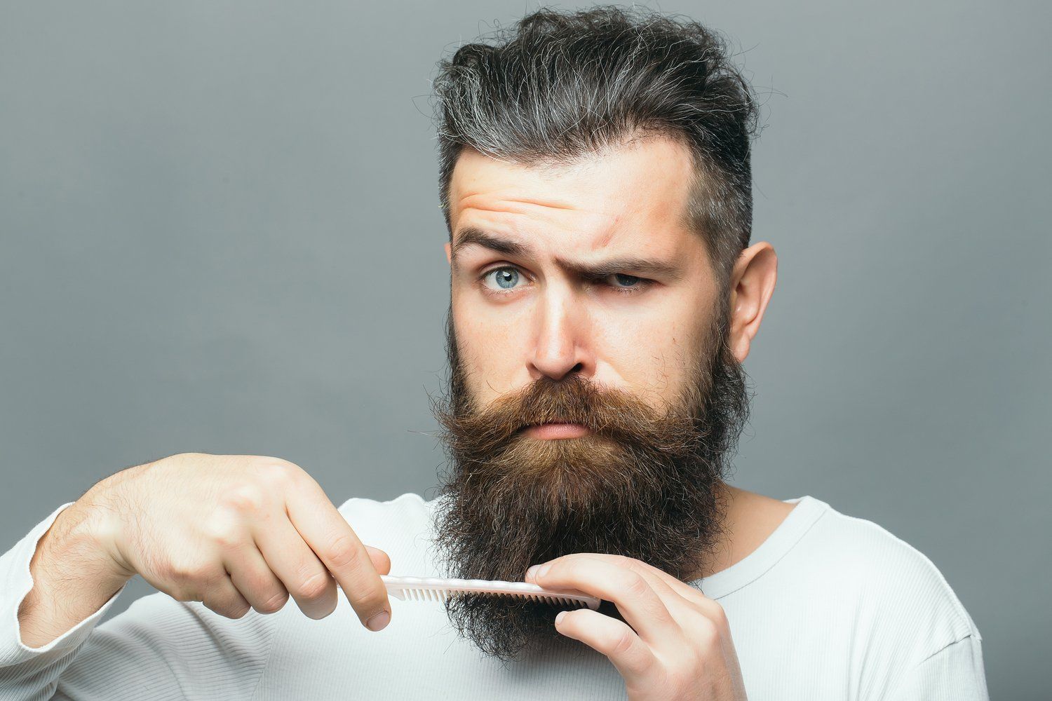 Hombre con barba poblada, peinándola con un peine pequeño, guiñando un ojo con un fondo neutro.