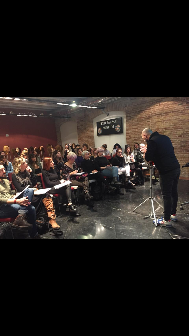 Un hombre calvo habla ante un público sentado en una sala con poca luz. La gente escucha atentamente; algunos toman notas.