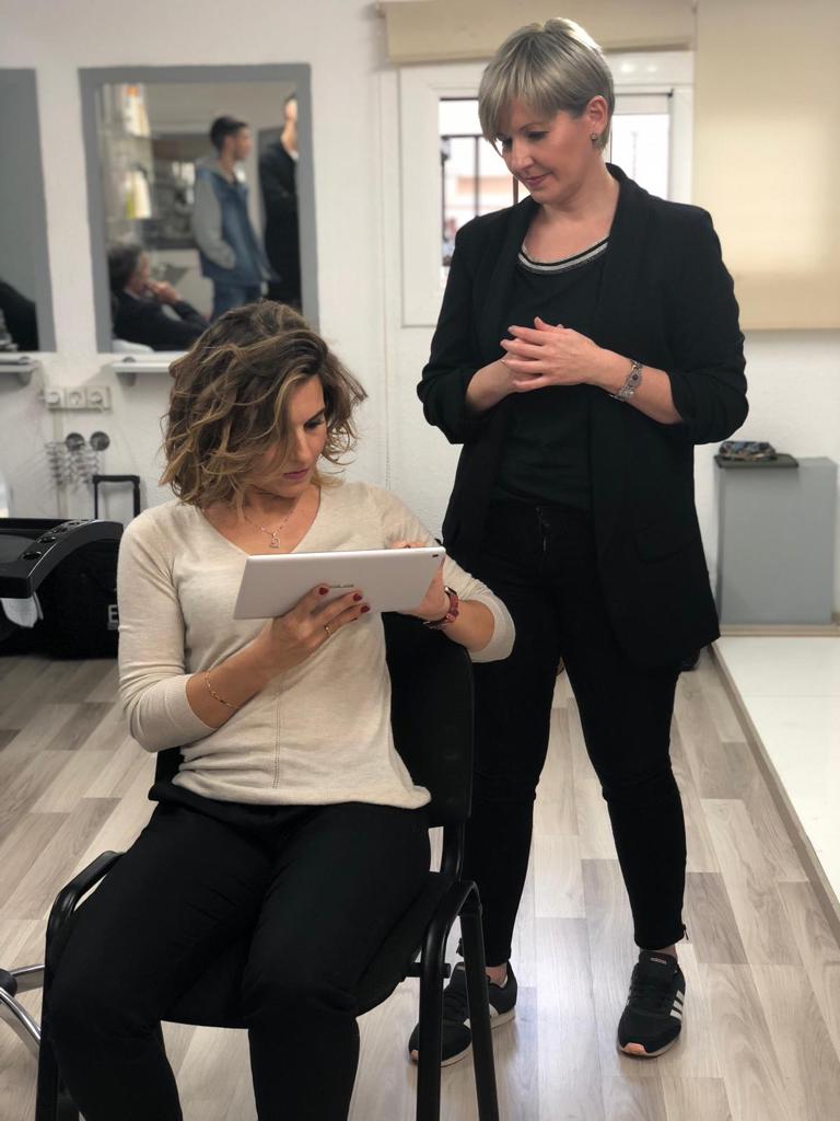 Mujer sentada revisando su tableta, otra mujer de pie observando. Salón de belleza, vestimenta informal.