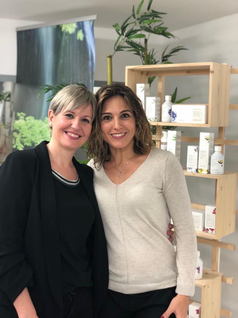 Dos mujeres sonrientes posan junto a productos para el cuidado de la piel en una habitación llena de luz.