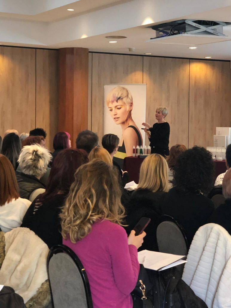 Taller de peluquería: Una mujer presenta su trabajo ante un público sentado. Un gran póster de una mujer rubia.