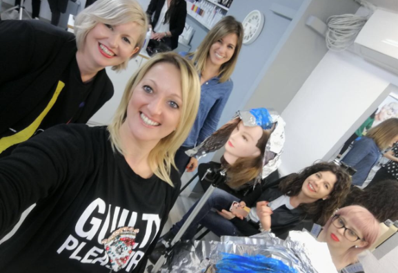 Mujeres en una peluquería, sonriendo a la cámara. Una rubia en primer plano, otras al fondo con maniquíes.