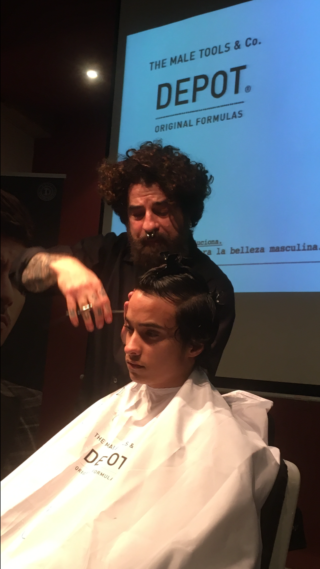Hombre corta el cabello de una persona con capa; evento con cartel de 