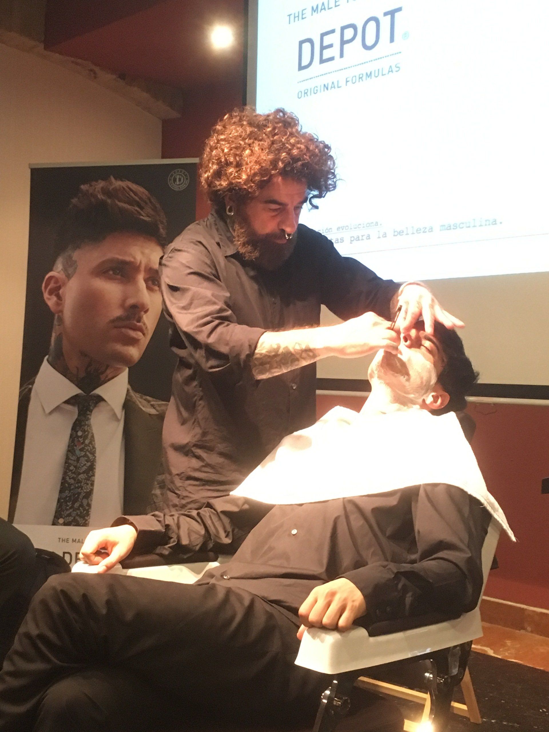 Un barbero afeita a un hombre en una silla. El barbero tiene el pelo rizado y tatuajes. Hay un cartel al fondo.