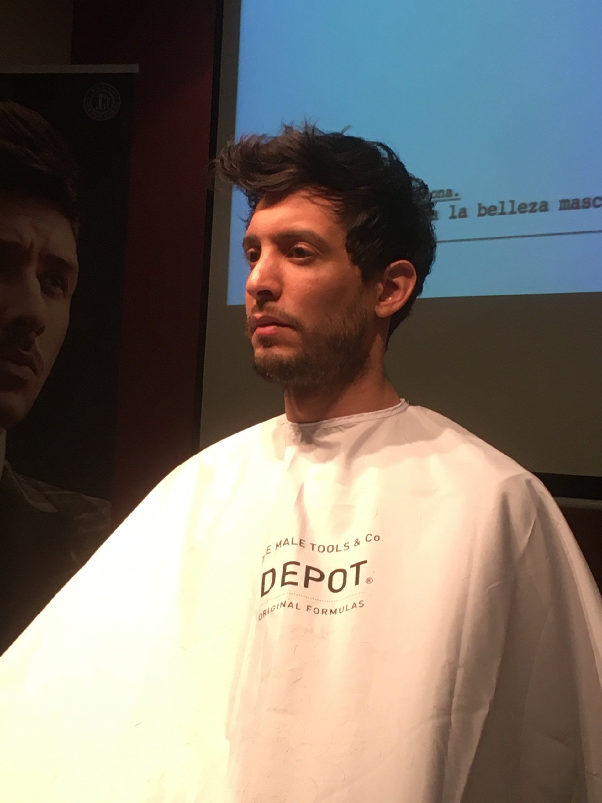 Hombre de pelo corto y oscuro con capa blanca de peluquería. Parece concentrado.