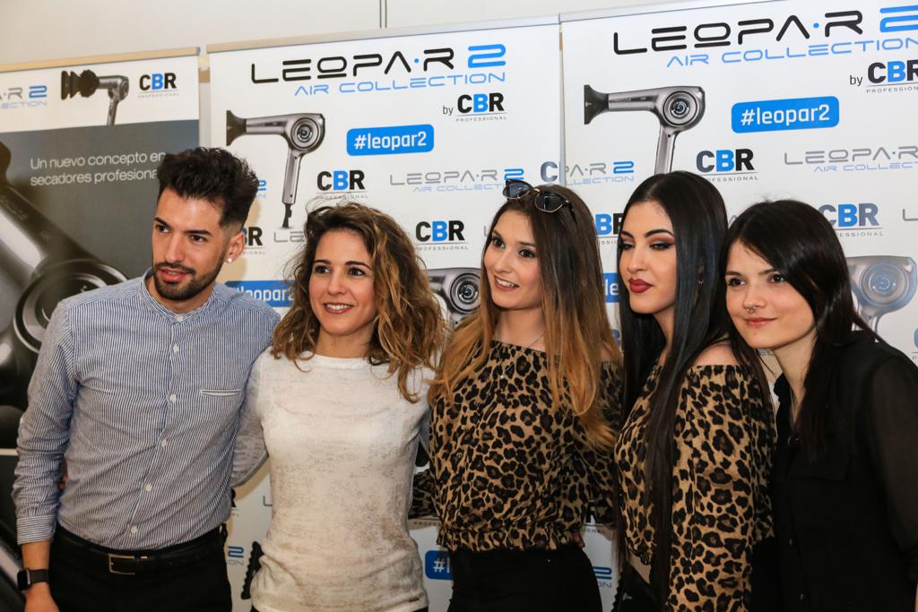 Grupo de cinco personas posando frente a una exhibición de secadores de pelo; todos sonrientes, con prendas variadas.