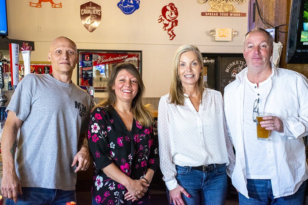 Four people pose together in a bar setting. From left to right: a man in a grey t-shirt, woman in floral top, and two others.