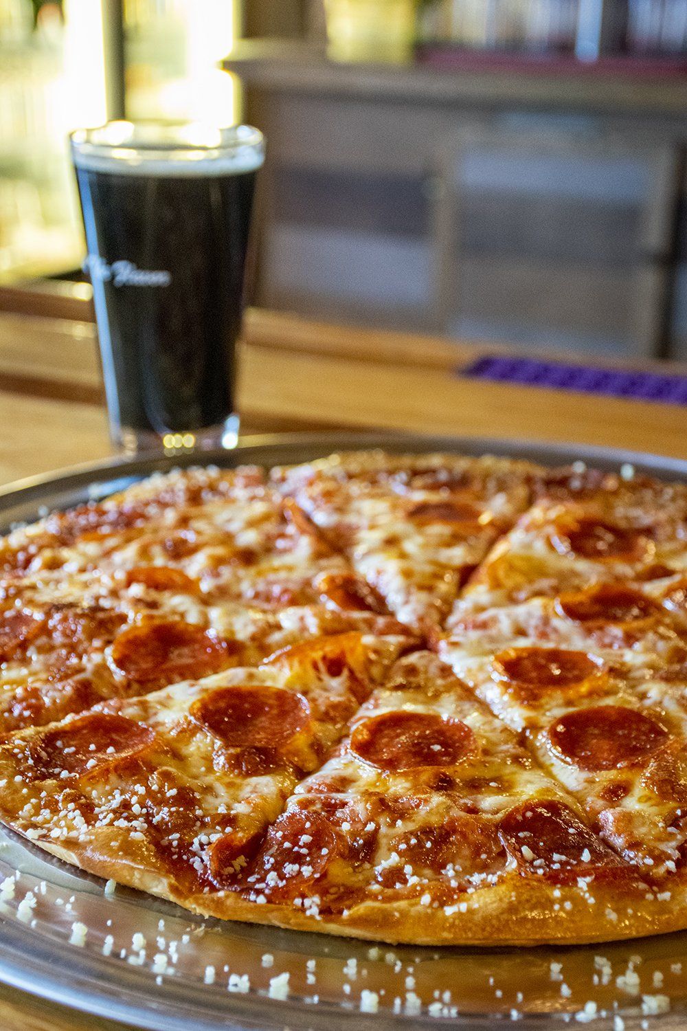 A freshly baked pepperoni pizza sits on a metal tray next to a pint glass filled with dark beer on a wooden bar top.