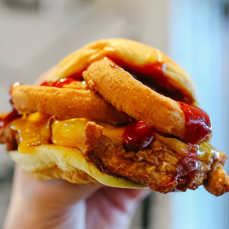 Hand holding a fried chicken sandwich with cheese, onion rings, and ketchup.