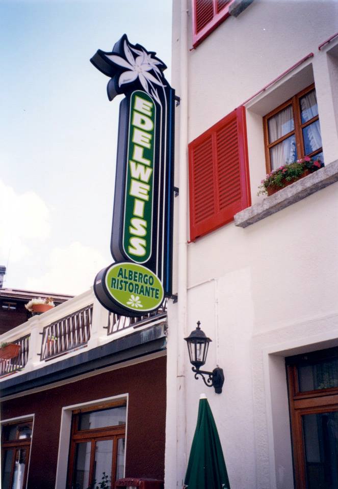 Un'insegna verticale verde e bianca dell'hotel e ristorante Edelweiss, montata sulla parete esterna bianca di un edificio.