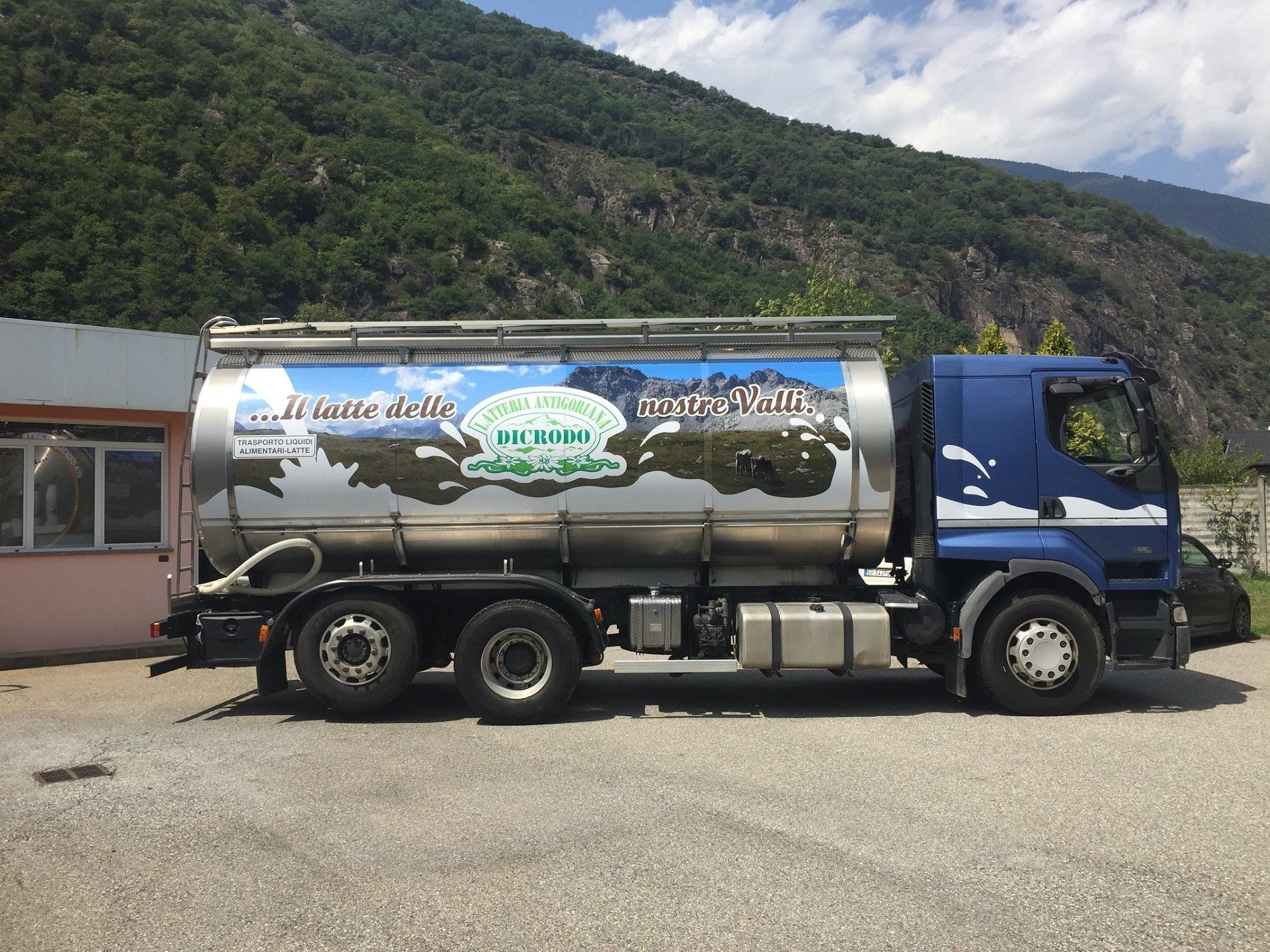 Un'autocisterna blu per il trasporto del latte parcheggiata all'aperto, con uno sfondo montuoso e una grafica paesaggistica decorativa.