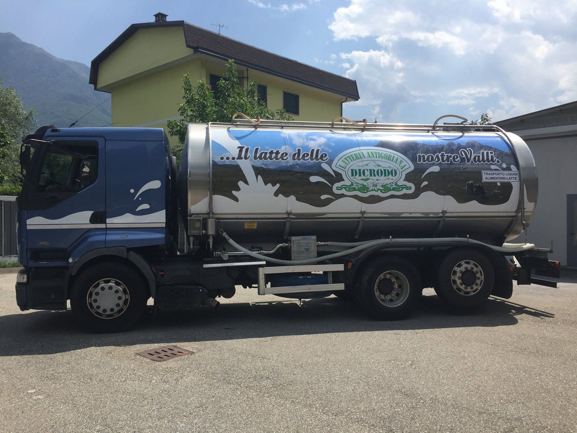 Un'autocisterna blu per il trasporto del latte, parcheggiata su un piazzale ghiaioso, con una grafica paesaggistica e il logo aziendale sul serbatoio cilindrico.