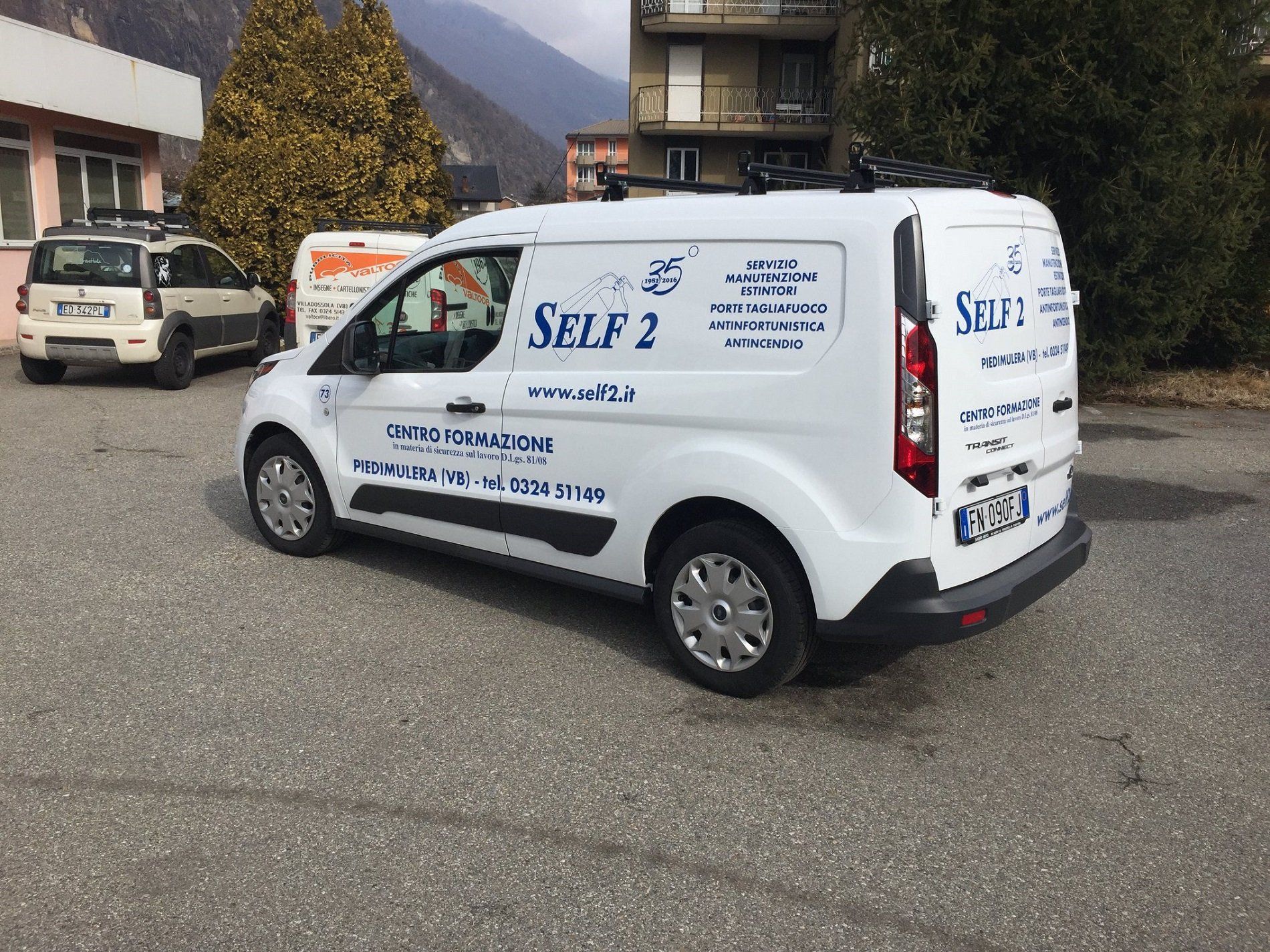 Un furgone bianco della compagnia Self 2 parcheggiato in un piazzale ghiaioso accanto a un'altra auto.