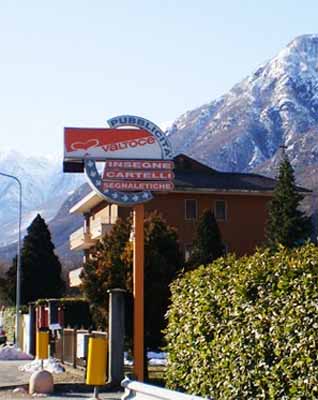 Un cartellone pubblicitario della Valtoce si erge su un palo di metallo, con una catena montuosa innevata sullo sfondo.