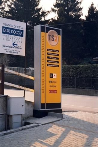 Un cartello giallo e nero di TS2, un'azienda di servizi IT, situato su un sentiero lastricato accanto all'ingresso di un edificio.