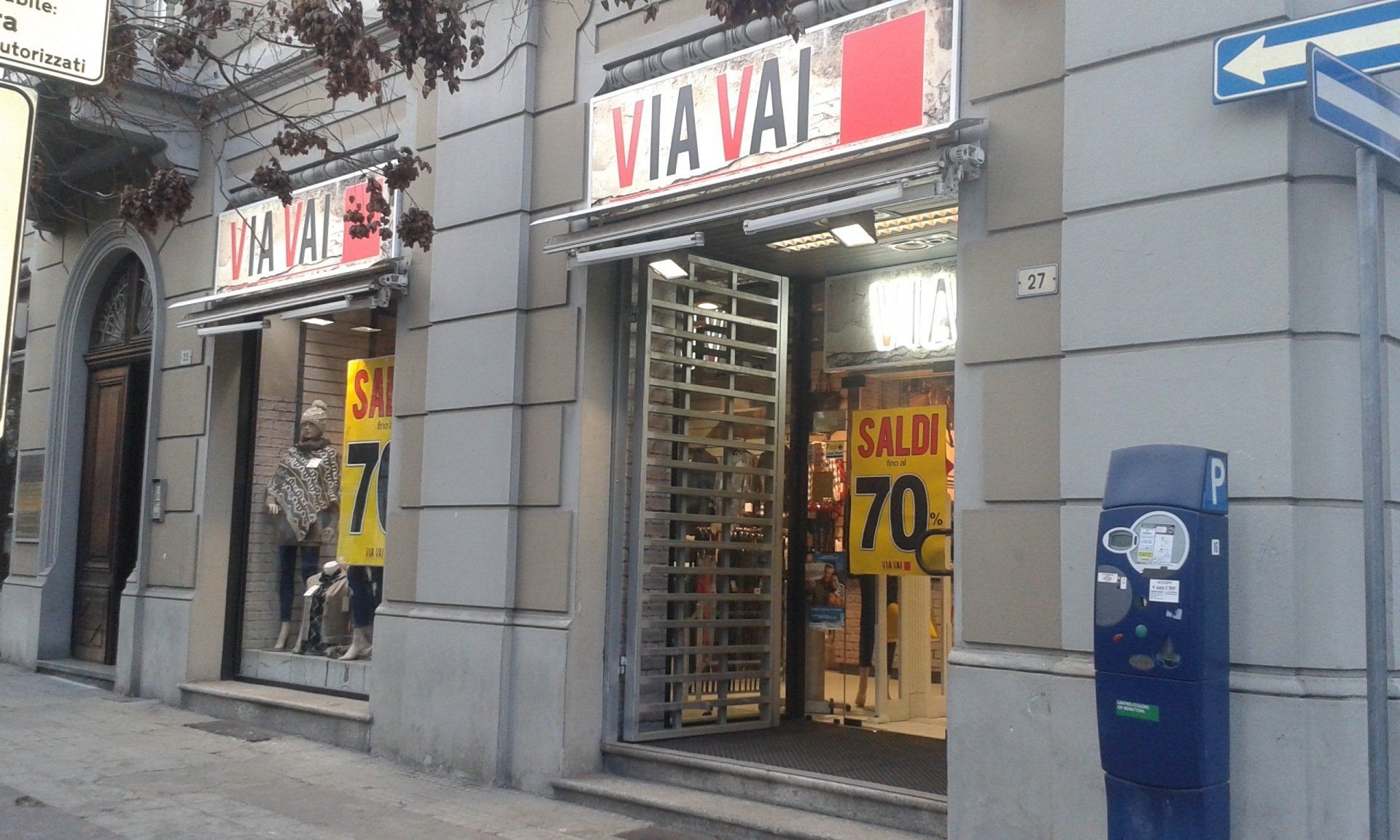 Un negozio con la scritta Via Vai, insegne “SALDI 70” in vetrina e un parchimetro sul marciapiede.