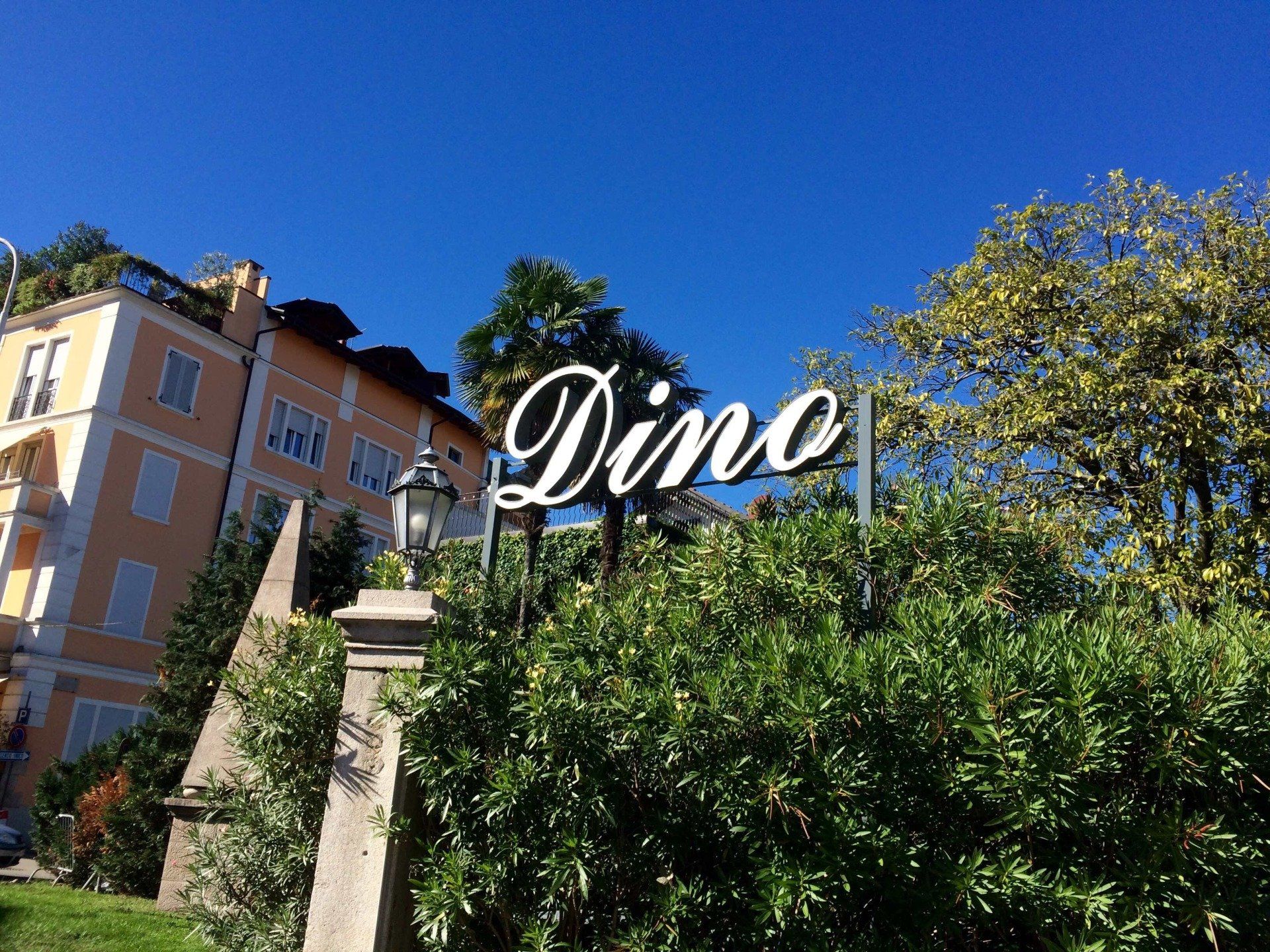In un giardino con alberi e un edificio color pesca, sullo sfondo di un cielo azzurro limpido, spicca un cartello bianco con la scritta