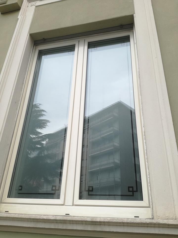 Una finestra bianca a doppio vetro incastonata in una parete beige, caratterizzata da un bordo geometrico decorativo nero nella parte inferiore dei vetri.