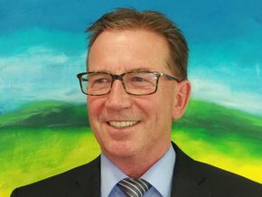 Mann mit Brille und Anzug lächelt vor einem farbenfrohen, gemalten Hintergrund.