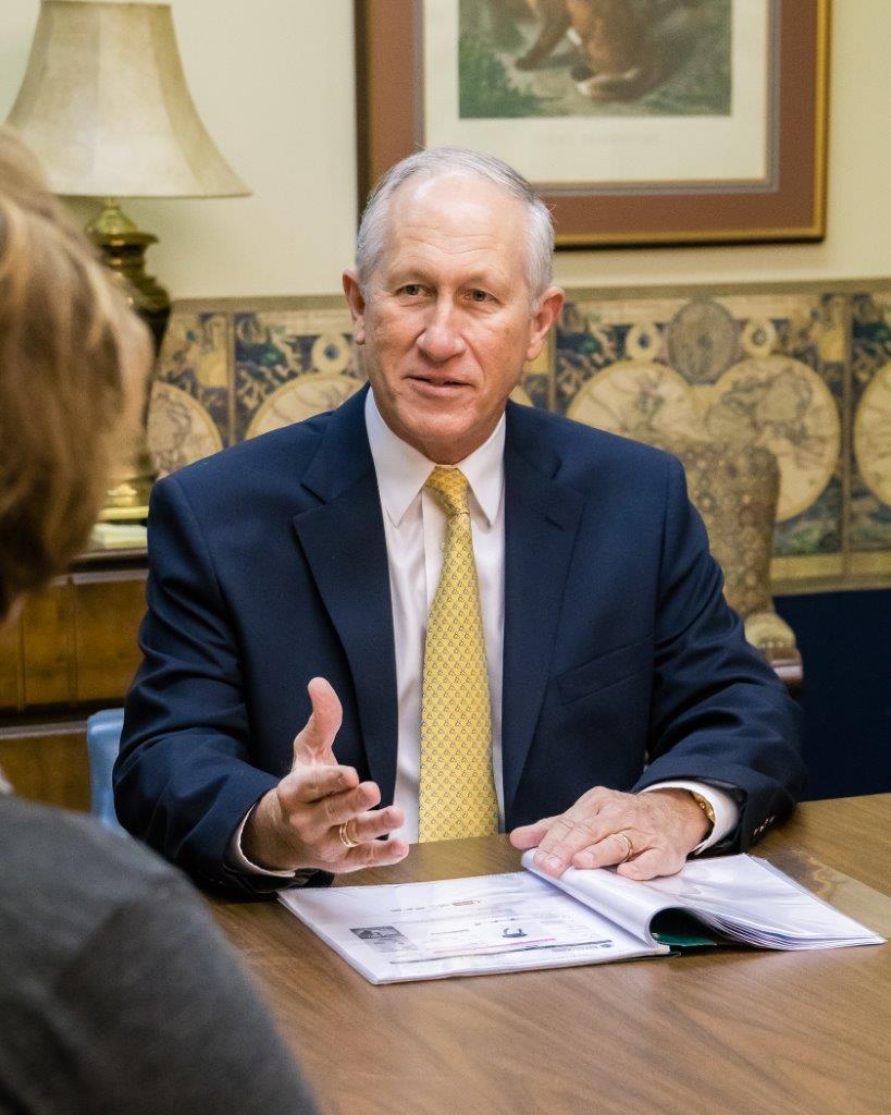 Man in blue suit with yellow tie gestures while speaking at a table; another person visible.