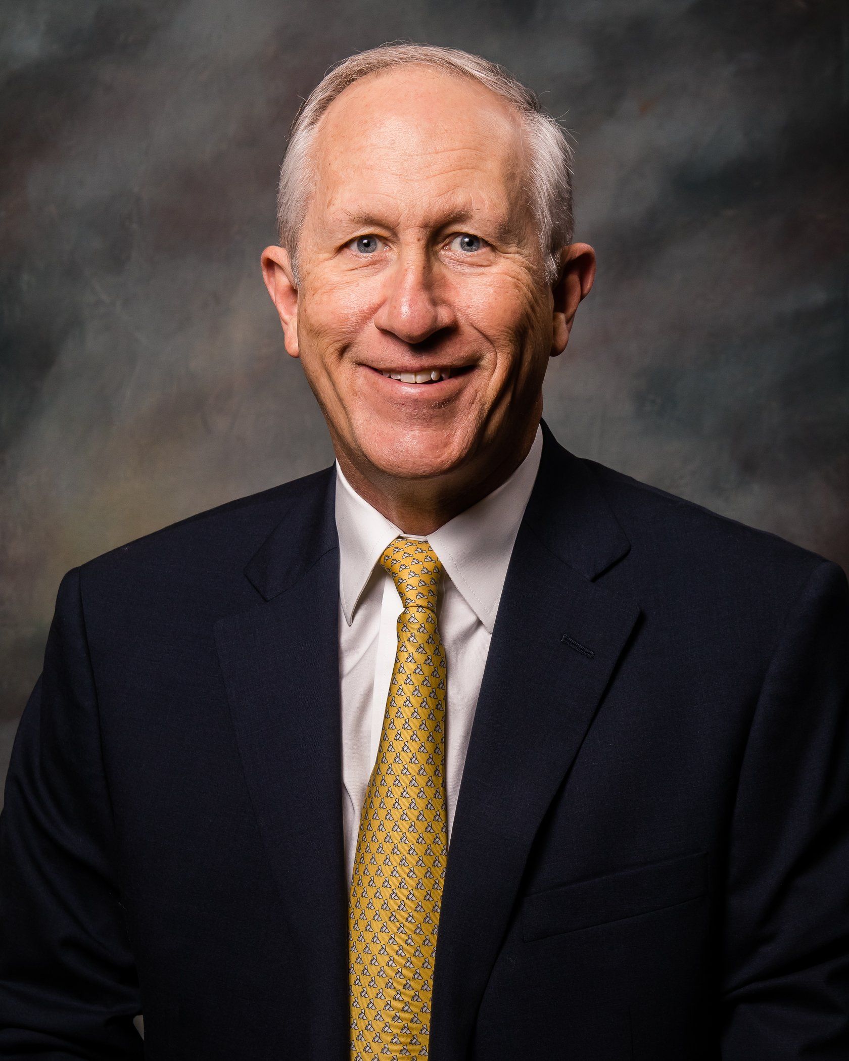 Man in suit smiles at the camera. He has fair skin, white hair, and a yellow patterned tie.