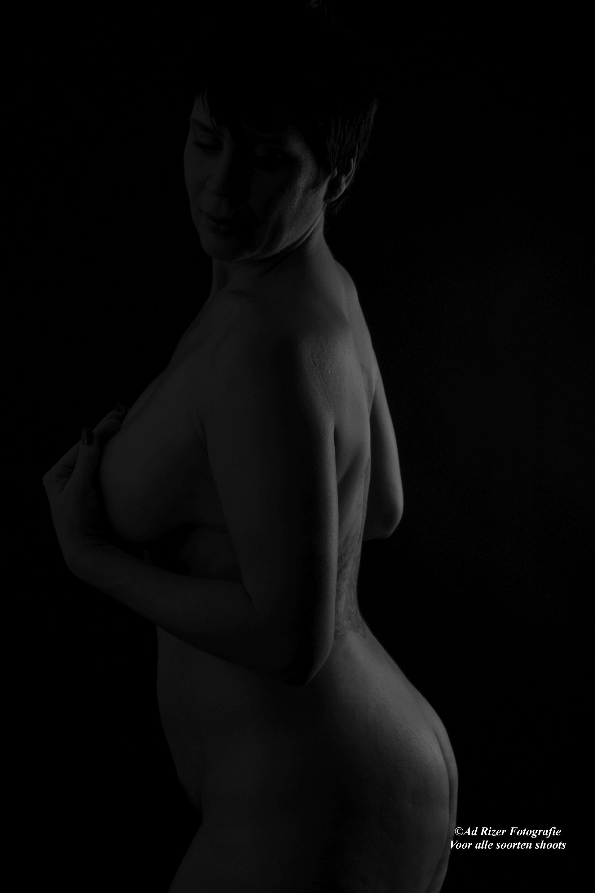 Zwart-witportret bij weinig licht van een naaktmodel, geposeerd in profiel tegen een donkere achtergrond.