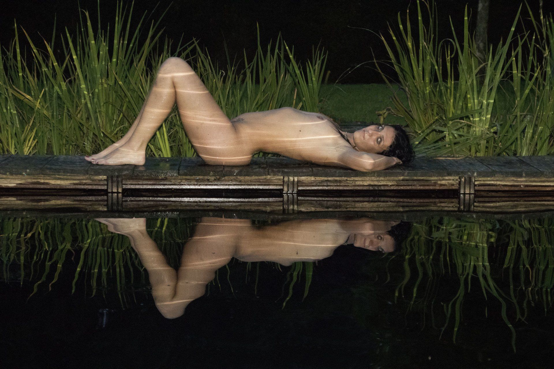 Naakt figuur liggend op een houten steiger 's nachts, weerspiegeld in het stille water met hoog gras op de achtergrond.