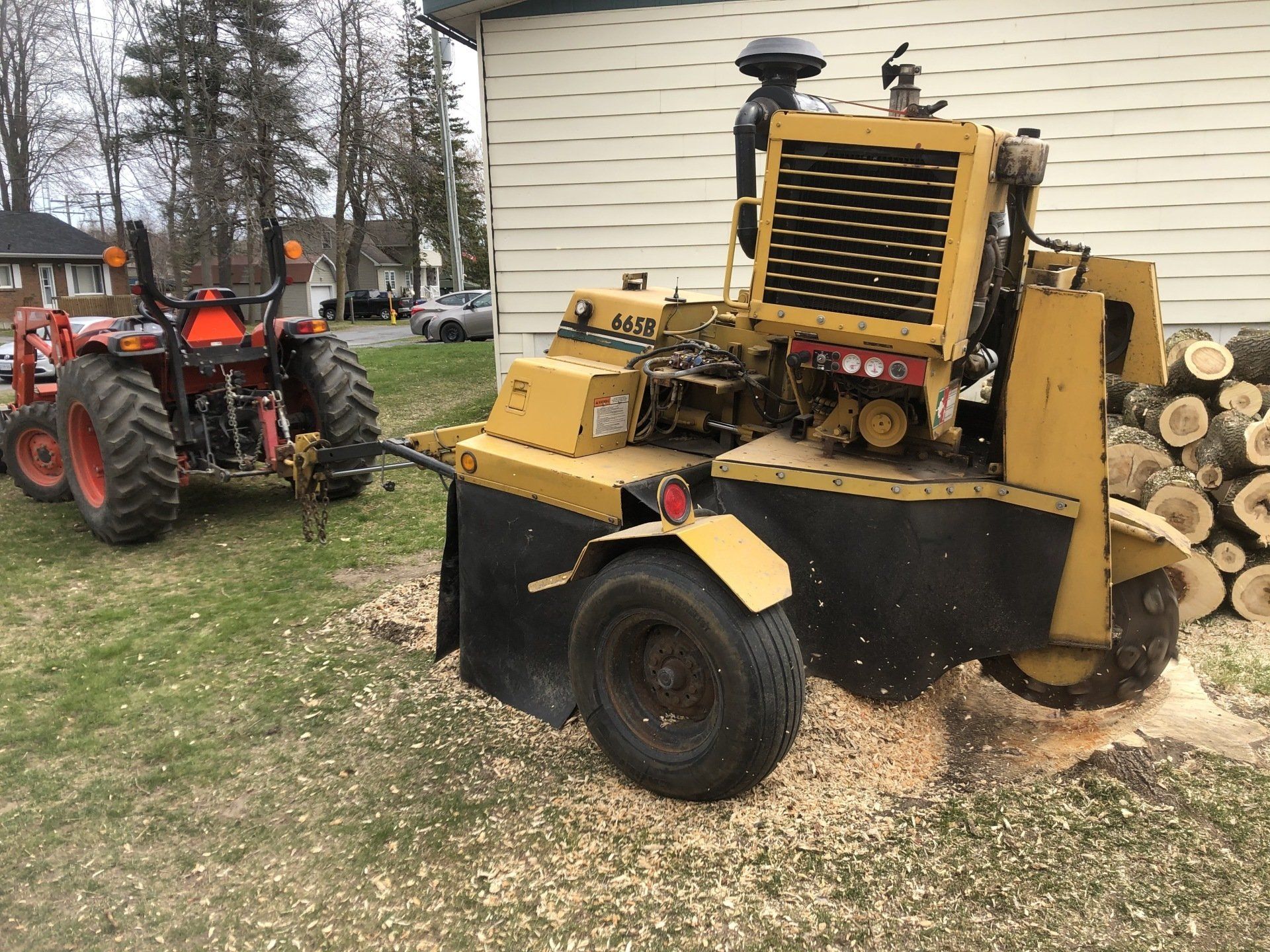 A tractor towing a yellow stump grinder on a grassy lawn next to a building.