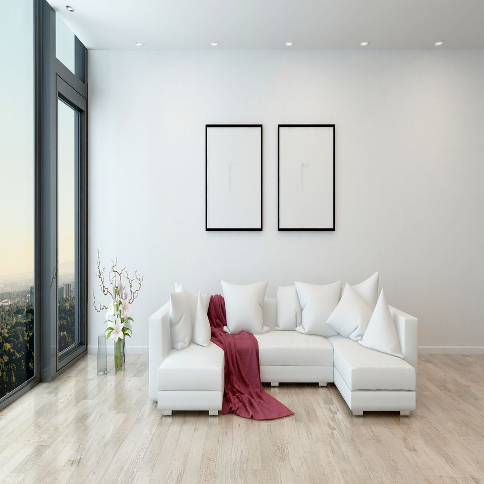 Soggiorno minimalista con divano angolare bianco, plaid rosso, ampia finestra e due stampe incorniciate alle pareti.