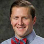 Man smiling, wearing a blue shirt and red bow tie, against a blurred backdrop.