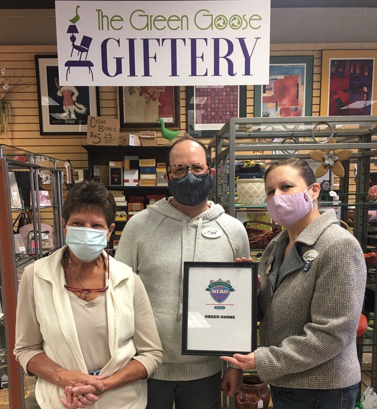 Three people in masks holding a framed award in a gift shop. Sign above says 