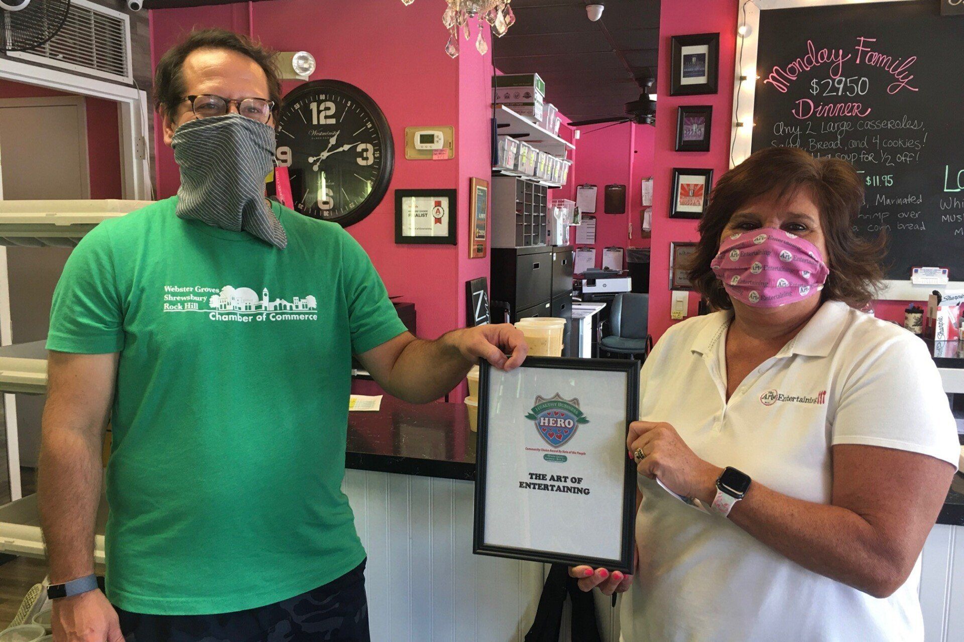 Two people, wearing masks, holding an award in a diner, with a pink wall.