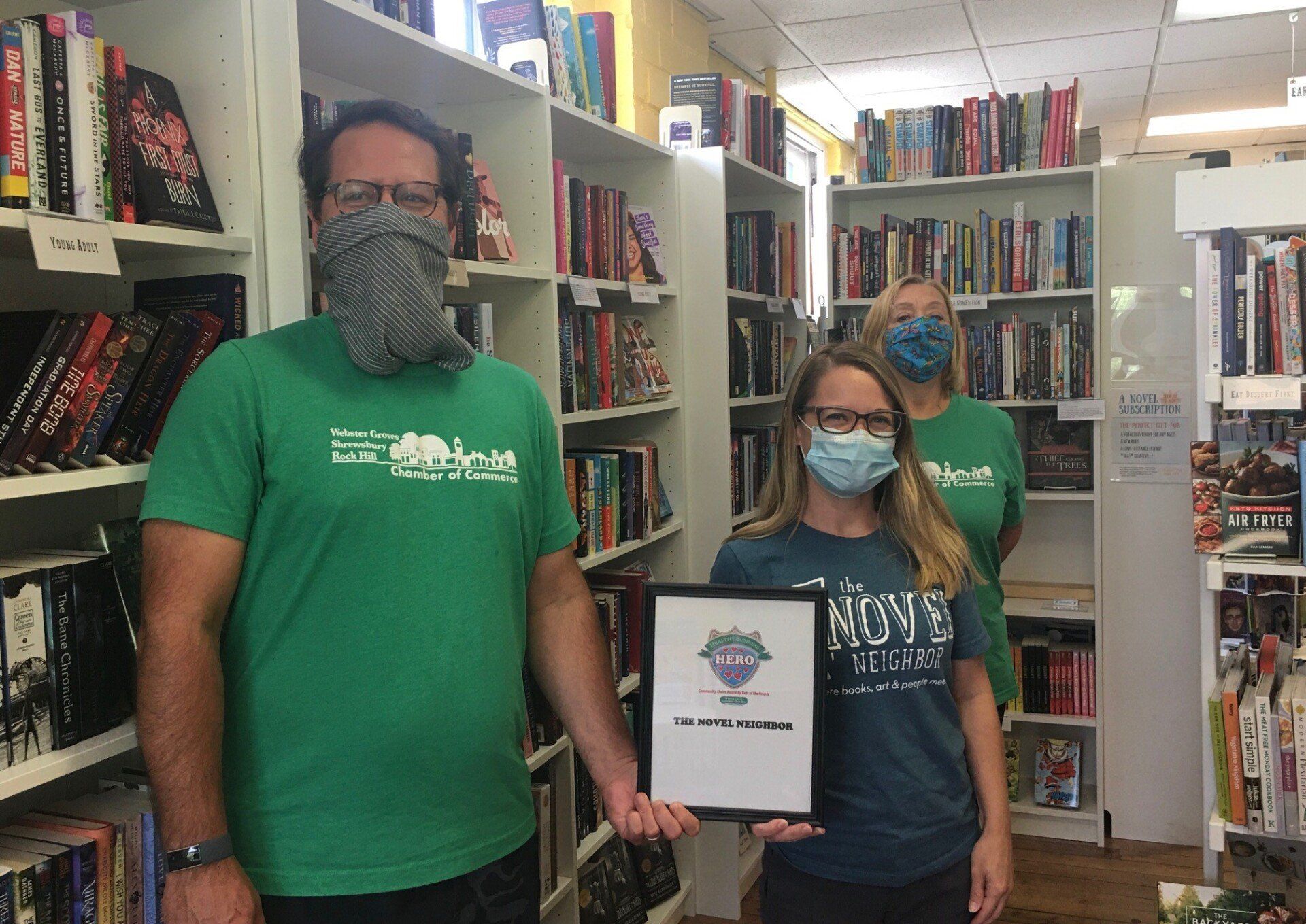Three people in a bookstore, masked, holding a framed award. Shelves of books in background.