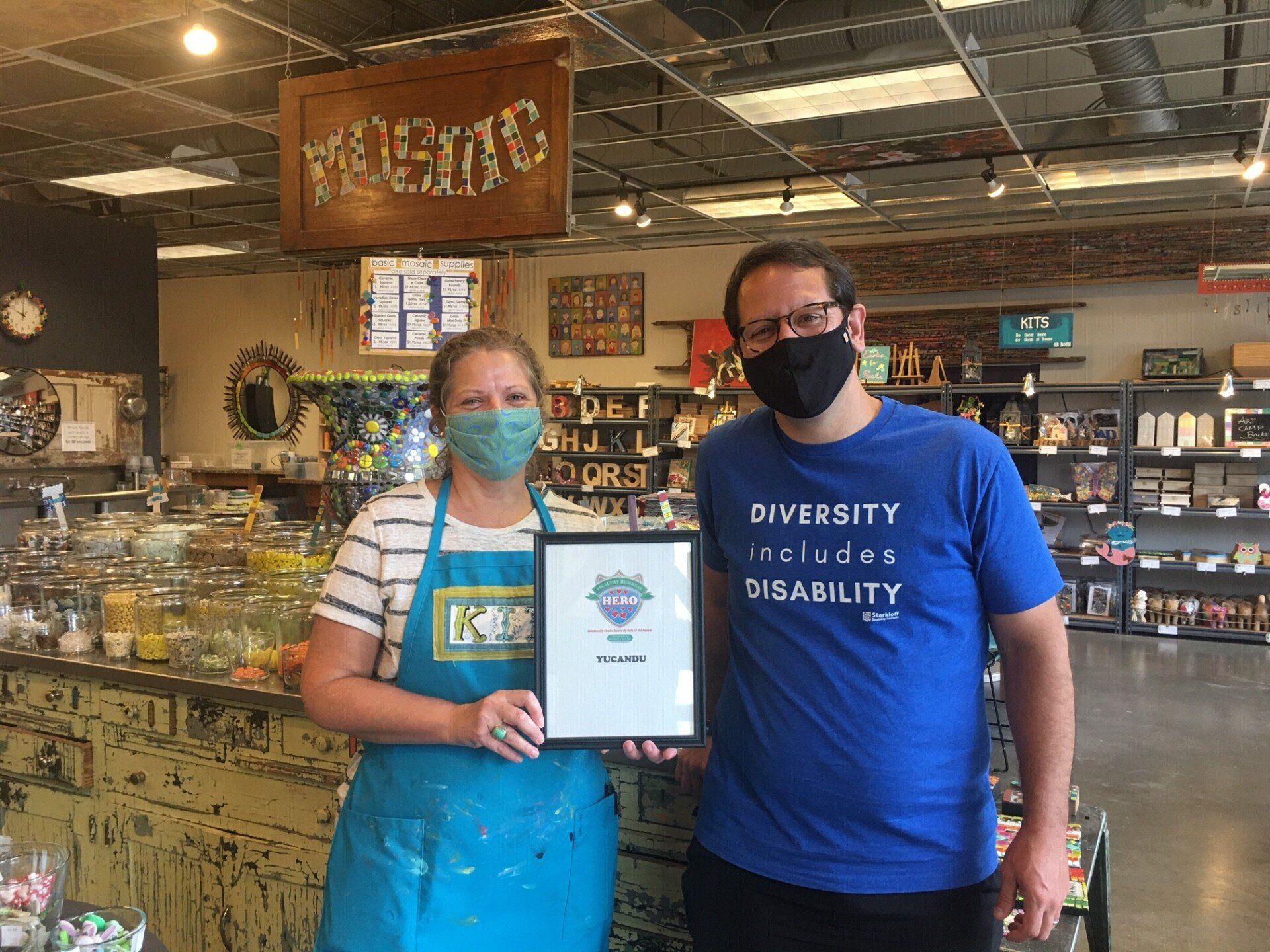 Two people holding a framed certificate in a candy store. One wears a mask and a blue 