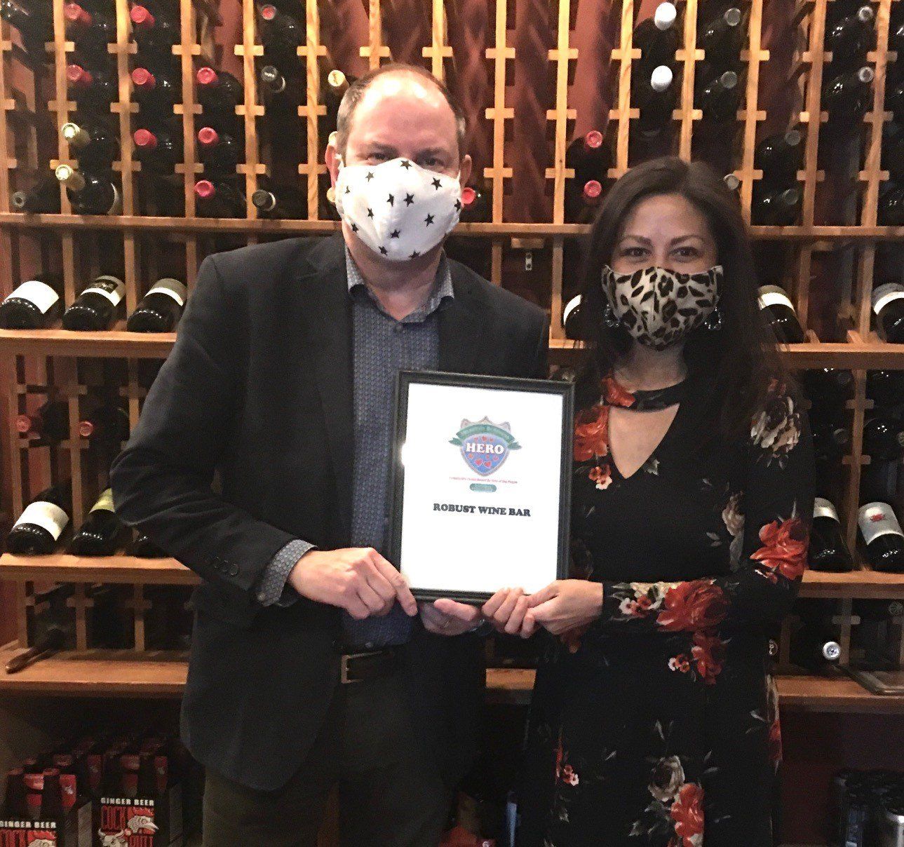 Two people with masks holding a framed award in a wine cellar.