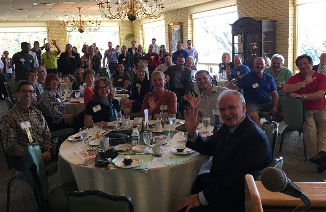 Large group of people at round tables in a brightly lit room, waving at the camera.