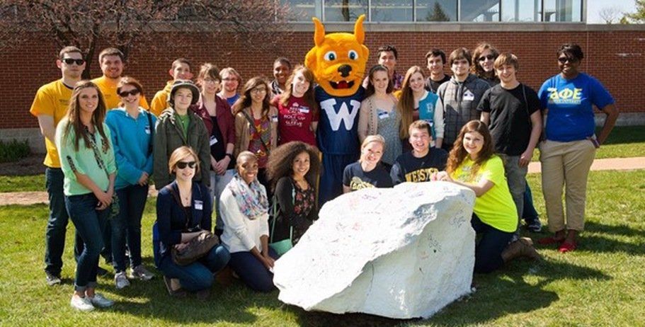Group of students and mascot pose near a painted rock on a grassy area outside a building.