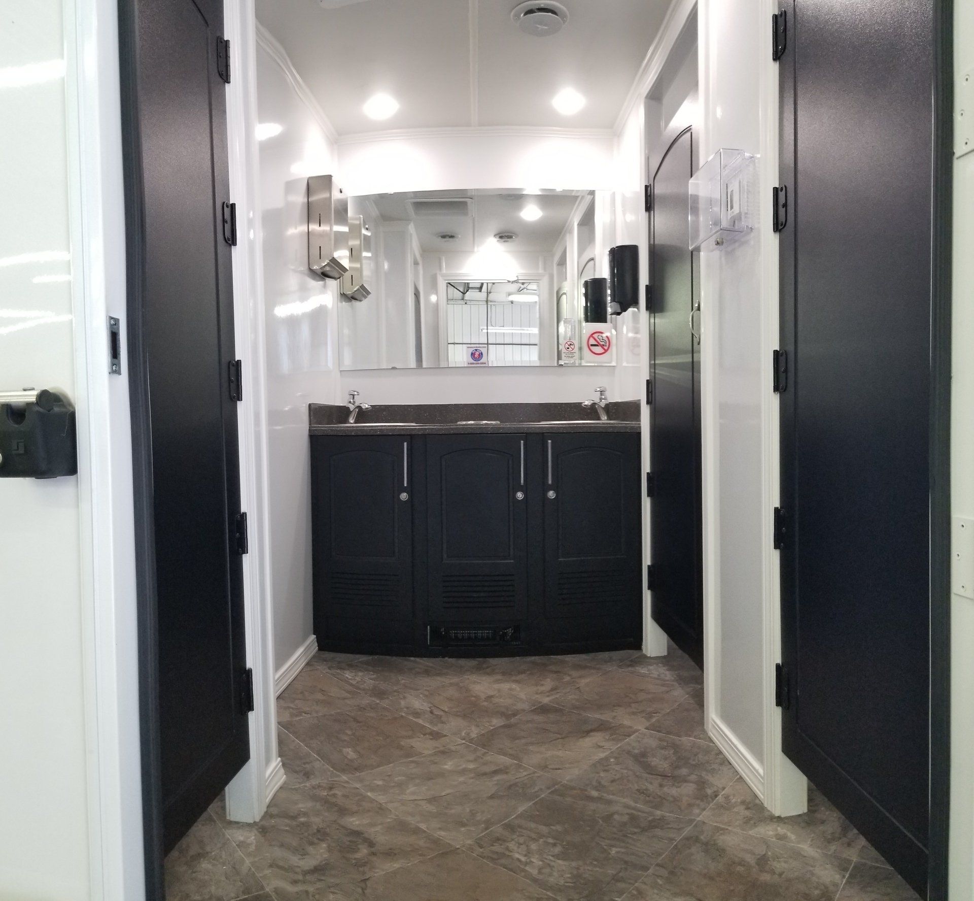 Interior of a portable restroom with two stalls flanking a central vanity with mirror and sink. Black and white.