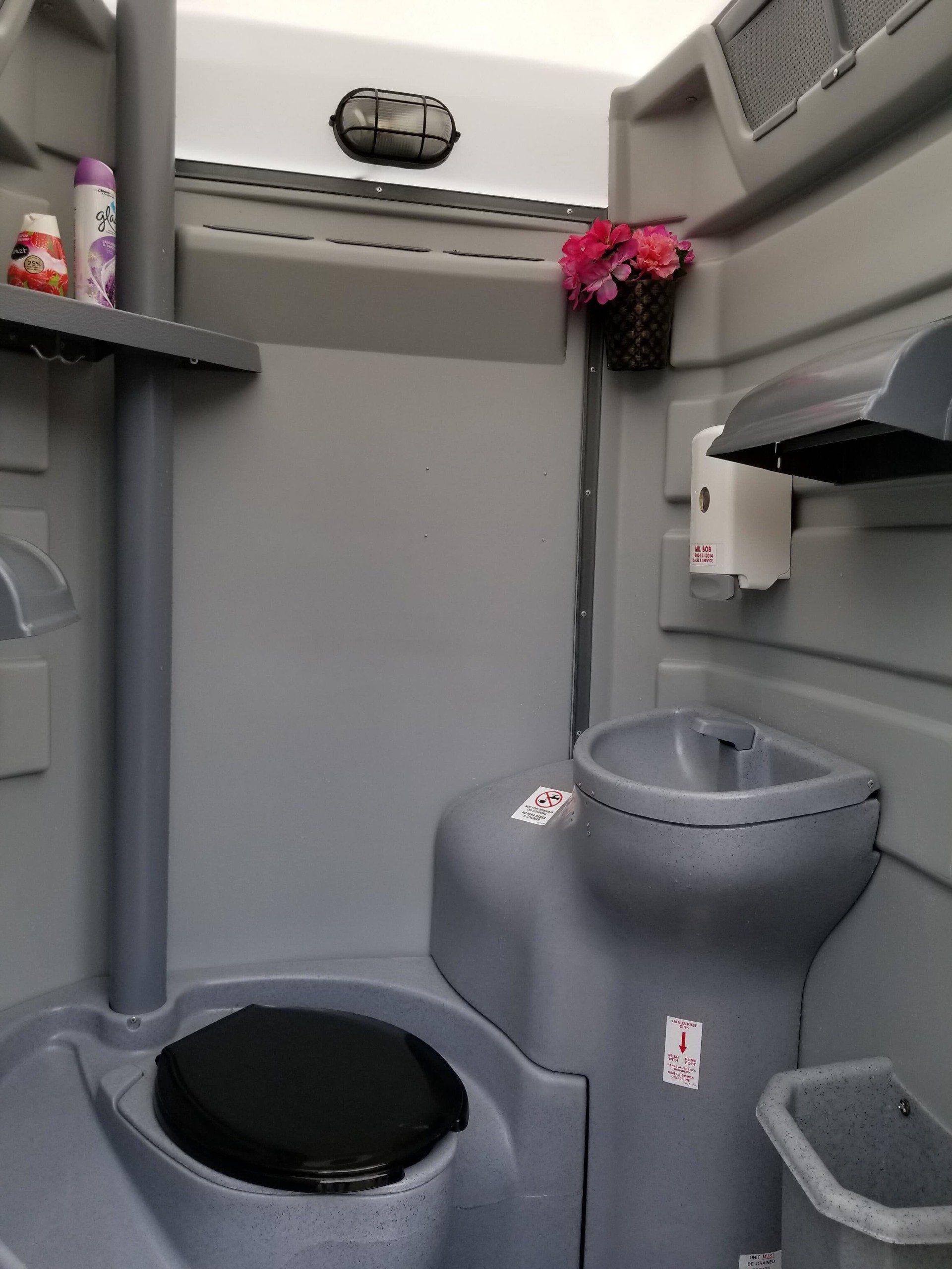 Interior of a portable restroom. Gray walls, toilet, sink, shelf with toiletries, and a small flower basket.
