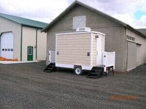 Mobile restroom trailer parked in front of a tan building with a barn door and a beige building with a white garage door.