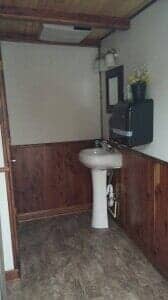 Bathroom interior with pedestal sink, mirror, soap dispenser, and wooden paneling.