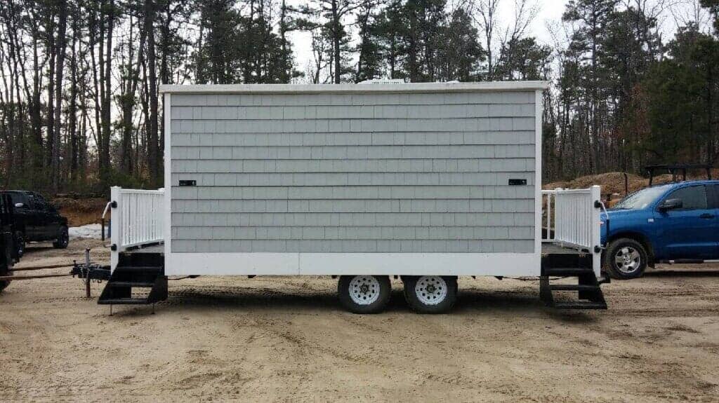 Gray-shingled portable restroom trailer with steps and white railing, parked on dirt.