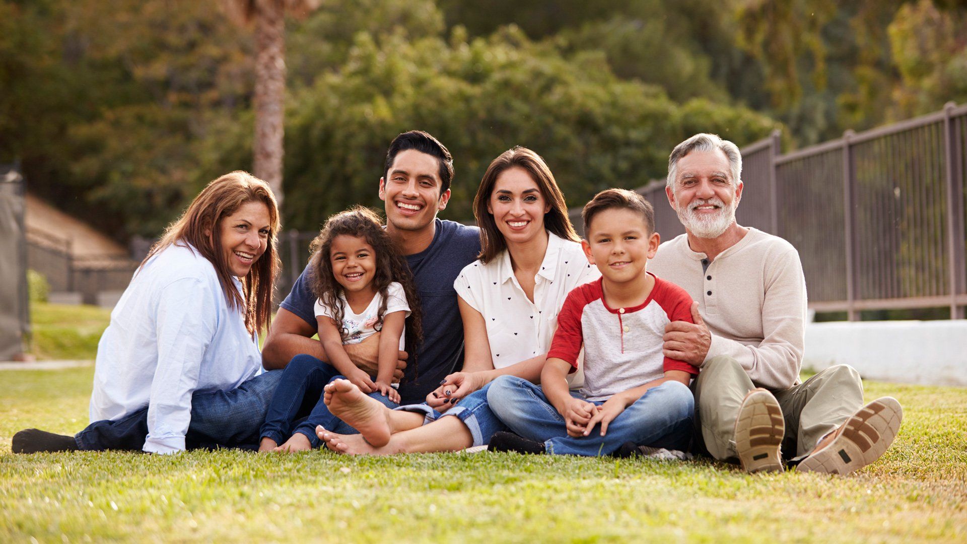 Family of six smiling and sitting together on the grass in a park.