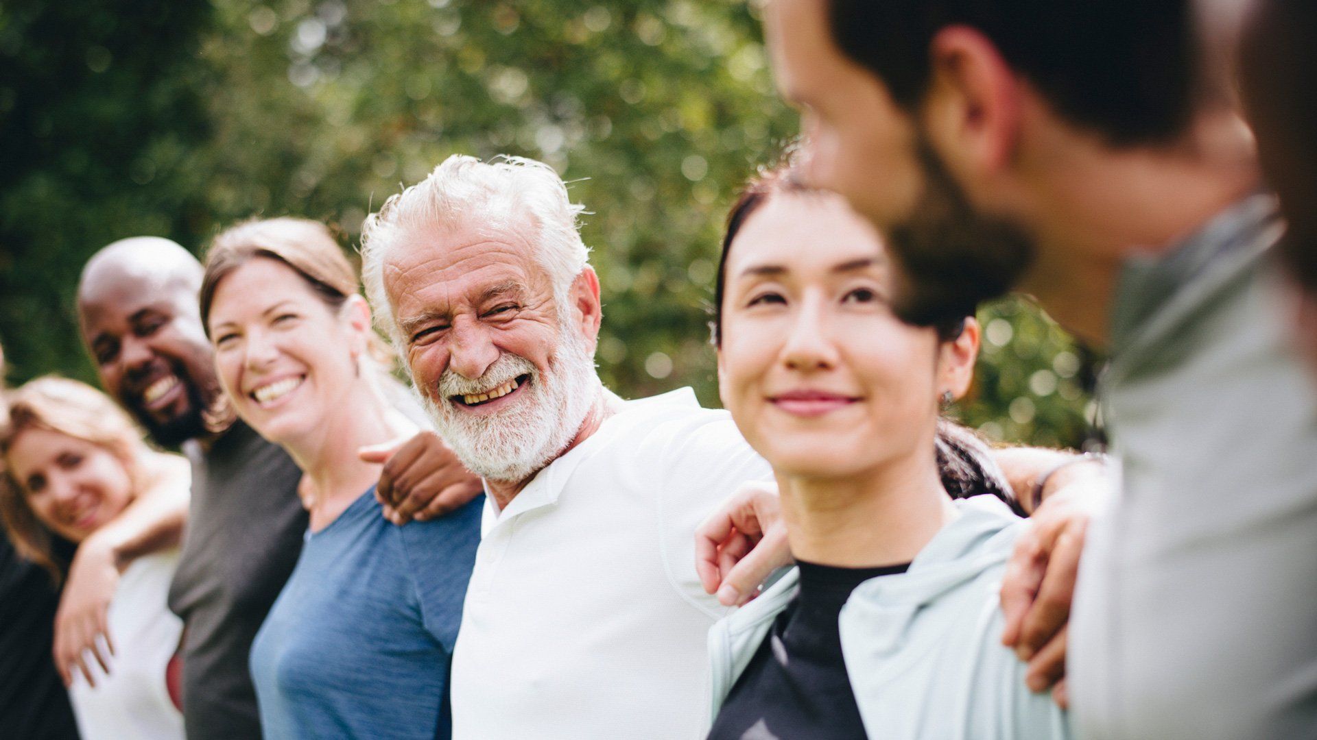 Group of diverse people with arms around each other, smiling, in a green outdoor setting.