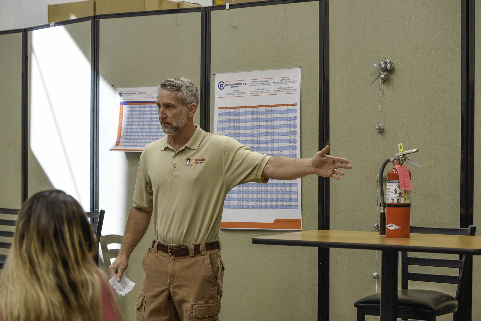 Man speaking in a room, gesturing beside a table with a fire extinguisher and posters behind him.
